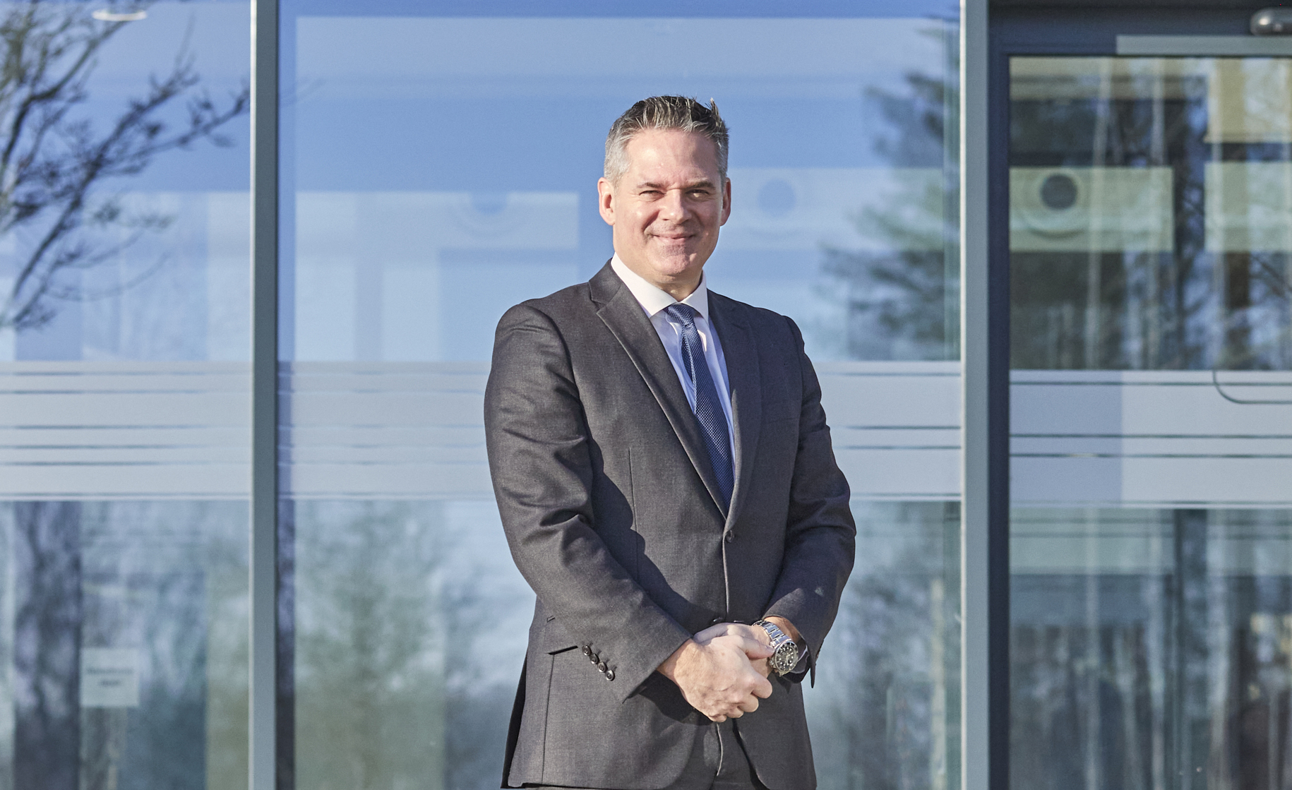 Managing Director, James Hindes, is standing in front of the Hoddesdon head office. He's wearing a business suit and it is a bright day.  Landscape crop.