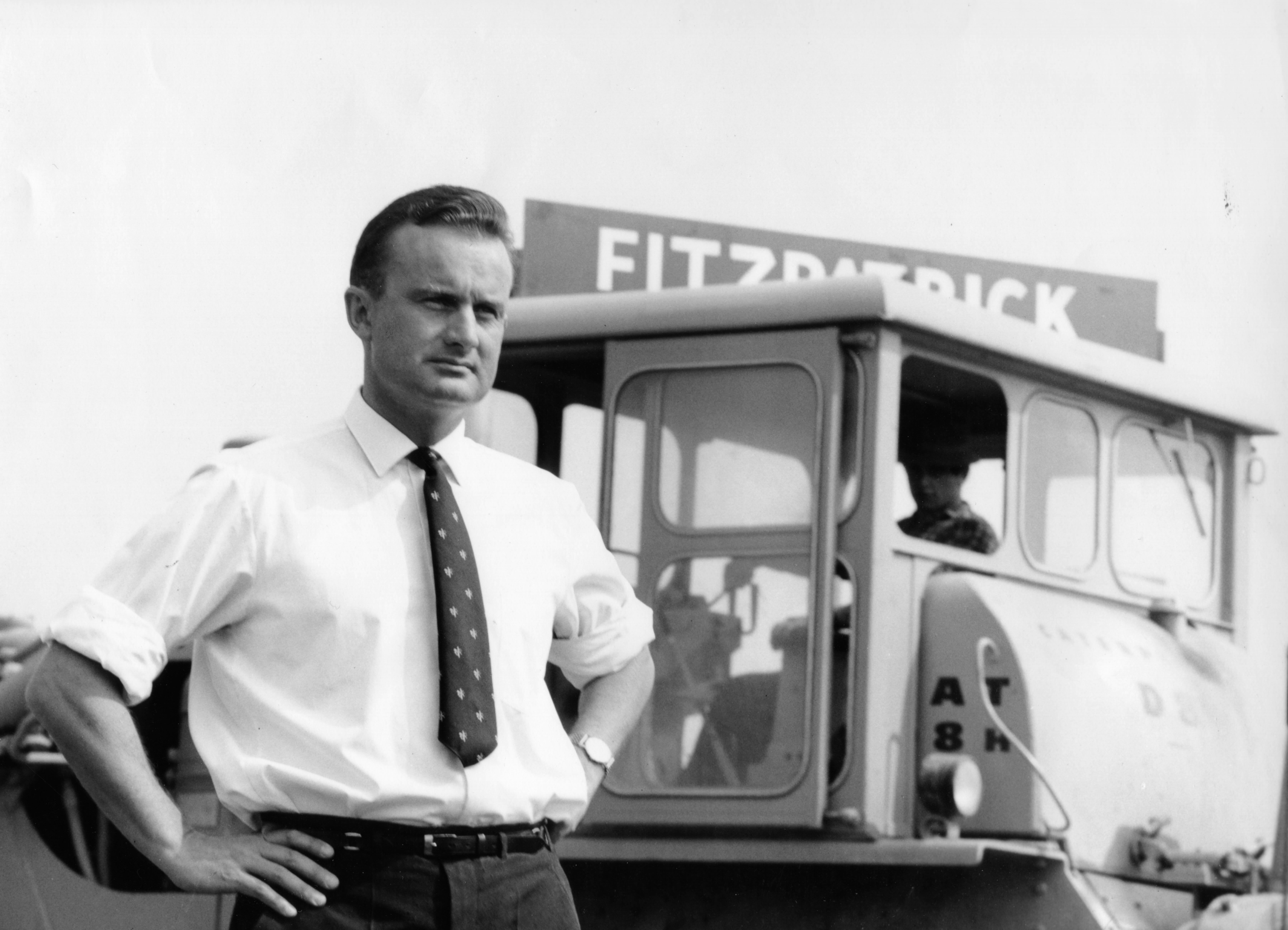 The original founder of the business is pictured in black and white, standing proudly with his hands on his hips, wearing a shirt and tie. In the background is a retro sign with the company name, Fitzpatrick, and a vintage digger. 