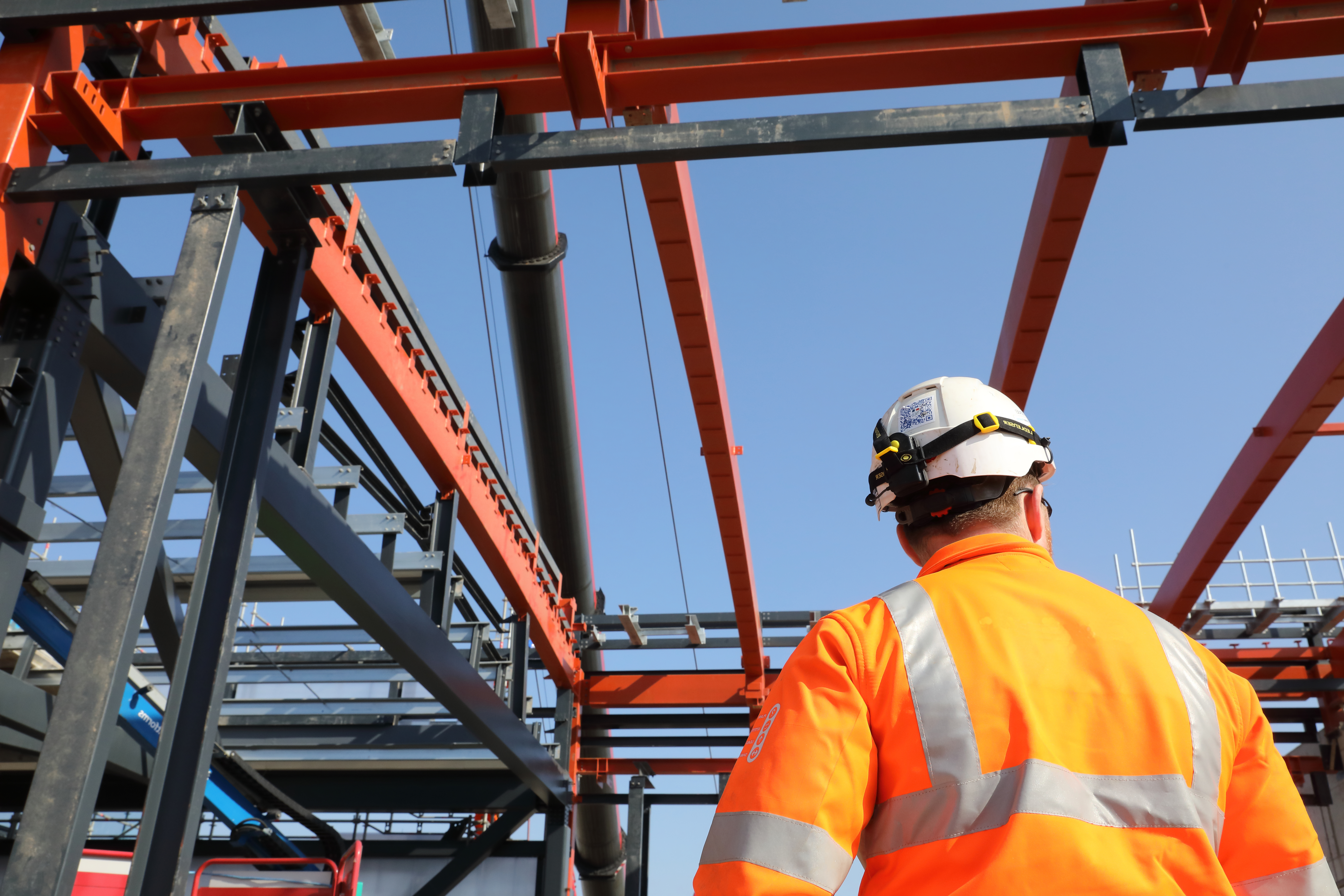 A man stands with his back to the camera. He is on site in high-vis orange clothes looking up at a red steel structure in a clear blue sky.